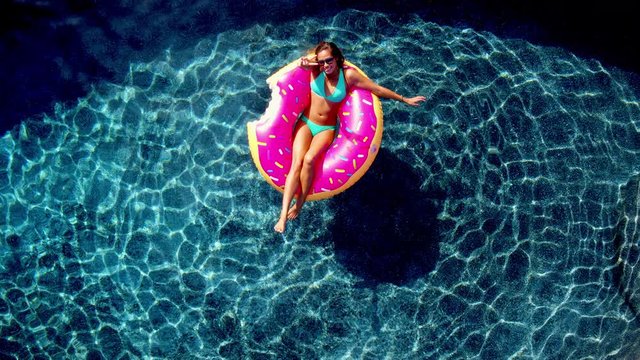 Young happy woman relaxing on inflatable pool toy in blue swimming pool on sunny day waiving at camera 