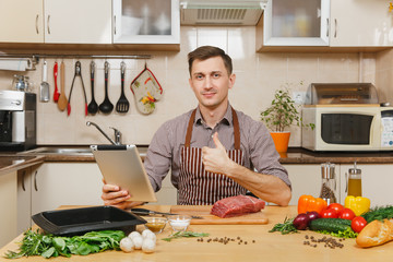 Young man in apron sitting at table with vegetables, looking for recipe in tablet, showing thumb...