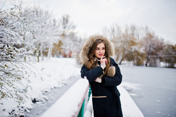 Beautiful brunette girl in winter warm clothing. Model on winter jacket against frozen lake at park.