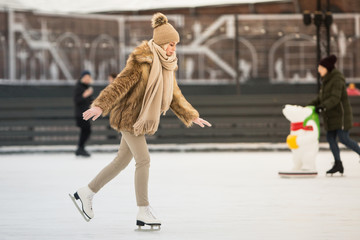 Full length portrait of young female with blonde hair in  fur coat, beige hat, scarf and trousers skating on ice rink, outdoors at winter /Weekends activities outdoor in cold weather/ - obrazy, fototapety, plakaty