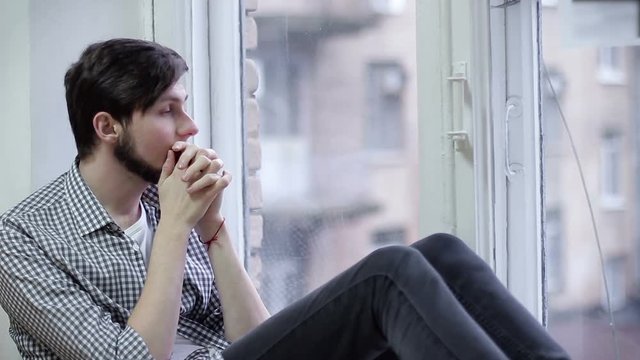 Sad guy sitting on the windowsill and looking out the window