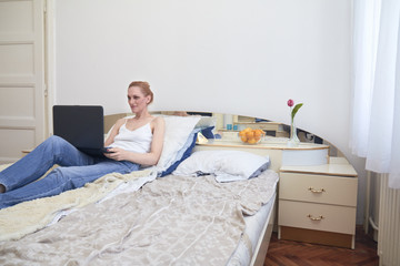 one woman, laying in bed smirking, using laptop. ordinary white room interior.