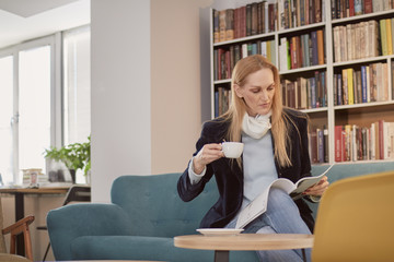 one woman, one person 40 years old, holding coffee cup, reading magazine, in library, book store, book shop, sitting in sofa. Shelf full of books behind (out of focus).