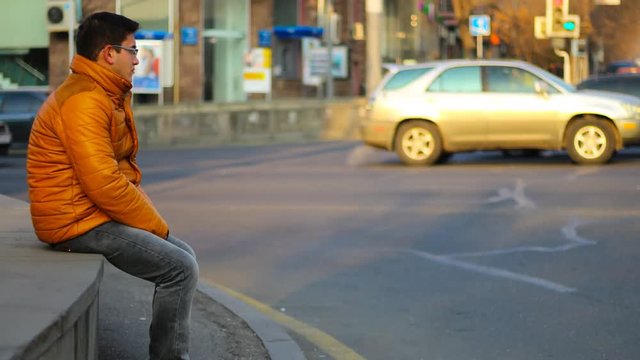 Sitting man in an orange jacket with thoughtful look, on the background of Time Lapse cars in city. 4K Time Lapse video.
