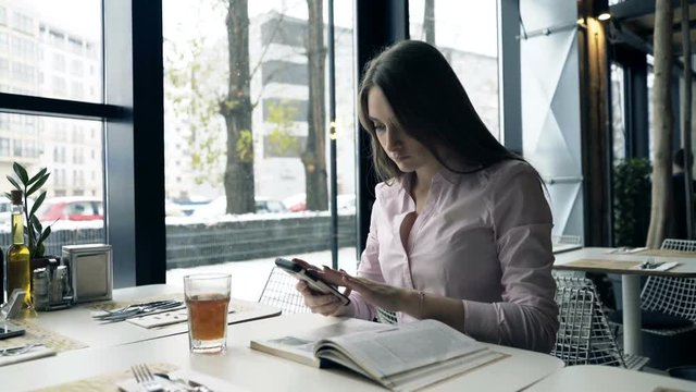 Young student girl with smartphone reading coursebook by table at cafe
