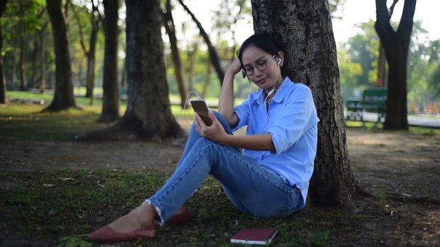 Asian woman listening to music from smartphone under the tree in the garden.