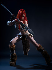 Beautiful red haired woman in stone age clothing with spear posing at camera on dark background