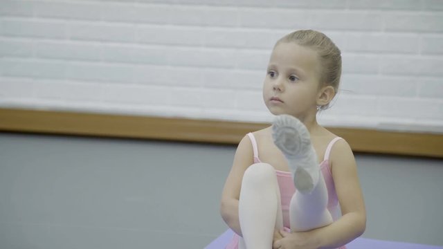 The portrait of girl who sits on the mat and stretch her legs in turn during the ballet club. Her knees of little are near the chest and straight one by one. The little dancer wears pink dress and gym