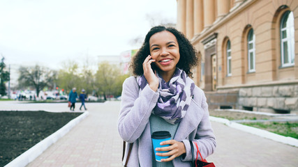 Attractive mixed race girl talking smartphone and drinking coffee walks in city street with bags. Young woman walking after shopping on mall sale