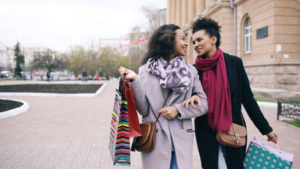 Two attractive mixed race women with shopping bags talking and walking down the street. Girlfriends have fun after visiting mall sale