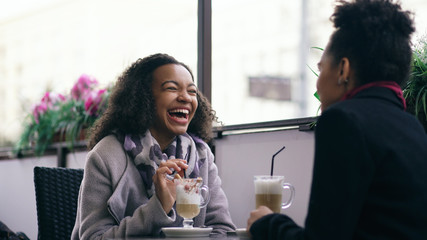 Two attractive mixed race women talking and drinking coffee in street cafe. Friends have fun after visiting mall sale