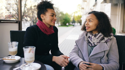 Two attractive mixed race women talking and drinking coffee in street cafe. Friends have fun after visiting mall sale