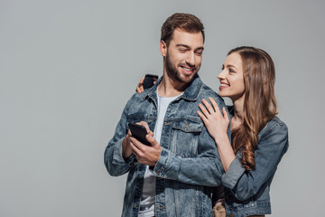 beautiful young couple in denim jackets using smartphones and smiling each other isolated on grey