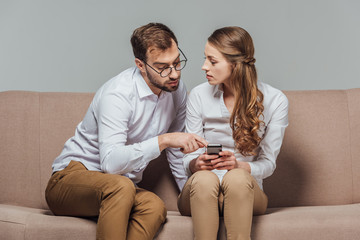 young woman holding smartphone and handsome man in eyeglasses pointing with finger while sitting on sofa isolated on grey