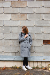 Portrait of tender smiling girl, fashion and stylish standing near rustic gray wall with a hole. Dressing autumn coat. Full-lenght