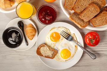 Plate with fried eggs and fork at the table with cup of black coffee, croissant, glass of orange juice, bowl with strawberry jam, bread on plate, fresh tomato