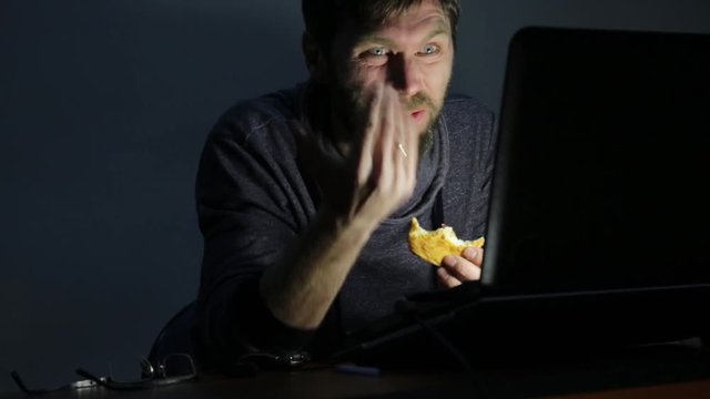 bearded man eating sandwich in front of a laptop, working and begins to upset
