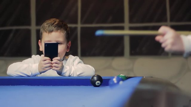 American billiard, nine-ball pool. Boy playing billiard, snooker while little brother takes a photo. Child shooting, hitting the ball. Kids in white play on blue table