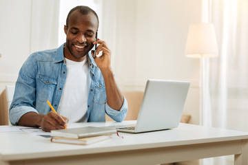 Listening. Attractive inspired young afro-american businessman smiling and talking over the phone and writing while sitting at the table and working on his laptop
