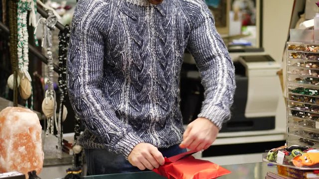 Male shopkeeper giftwrapping product in his store for a customer, putting it inside giftwrap bag and applying ribbon