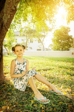 Summer education background: cute little girl sitting on grass and writing in notebook under the tree in warm sun rays. House and trees in background. Homeschooling in tropics concept. Text space