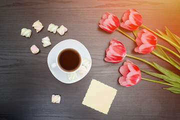Fototapeta na wymiar Top view on mug of coffee, marshmallows, blank postcard and pink tulips. Black background.