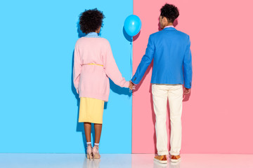Rear view of young couple holding hands with air balloon on pink and blue background