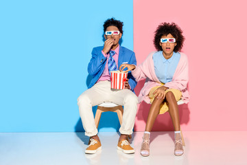 Obraz premium young african american couple sitting on chairs and eating popcorn against pink and blue wall