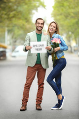 Young man holding paper with text SHE SAID YES and his happy beloved on engagement day outdoors