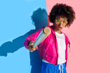 Young angry african american girl pointing in camera with baseball bat on pink and blue background