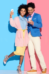 Obraz premium Young african amercian girl taking selfie and hugging stylish guy who looks at his phone on pink and blue background
