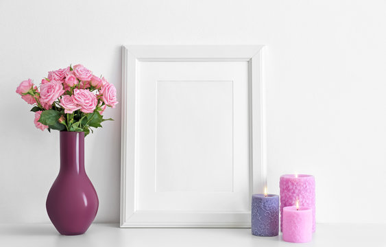 Empty frame, pink roses and candles on table near white wall