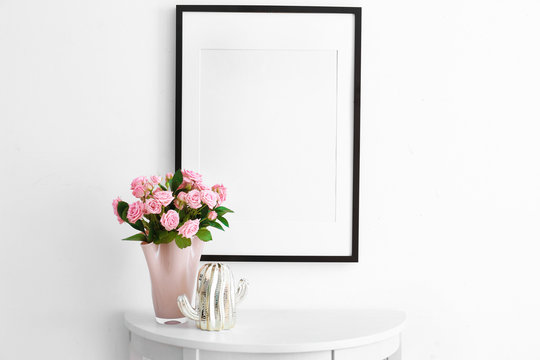 Empty frame, pink roses and decorative cactus statuette on table near white wall