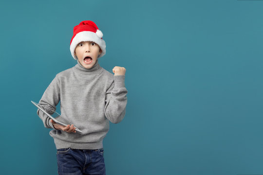Cute boy in Christmas hat standing with tablet and celebrating with fist up.