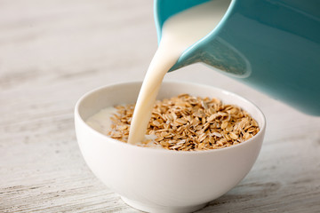 Milk pouring into bowl with oatmeal flakes on wooden background