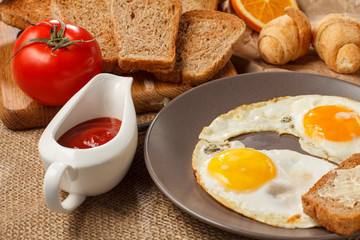 Plate with fried eggs, toast with butter, croissants, bread on wooden cutting board, fresh tomato and ketchup in a sauce boat