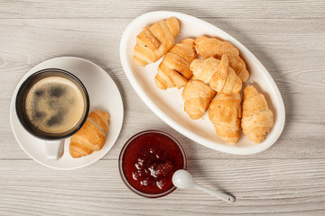 Cup of black coffee with saucer, fresh croissants on white porcelain dish, strawberry jam in bowls