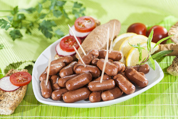 Chicken & Beef cocktail sausage with cherry tomato radish and lemon with green herb with toast bread on green red and bamboo background  with white and green plate