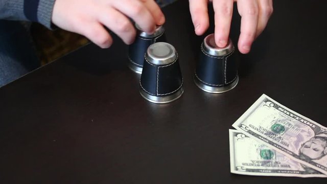 Thimblerig. A man hides a pea under one of the glasses, moves them on the table. The player points to the glass, but does not guess, and gives the win to the presenter. Cups and balls trick.