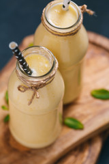 The yellow fruit smoothie in the glass bottles with straw on the wooden decorative cutting boards on the black table.