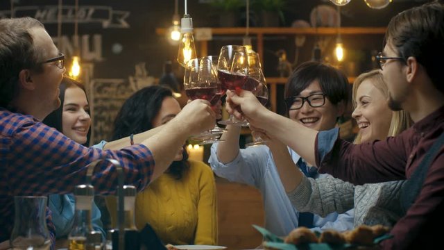 Diverse Group of Friends Celebrate with a Toast and Clink Raised Wine Glasses in Celebration. Beautiful Young People Have Fun in the Stylish Bar/ Restaurant.