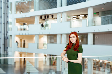 Fototapeta na wymiar Business portrait of the beautiful red-haired girl in a green dress against the background of office.
