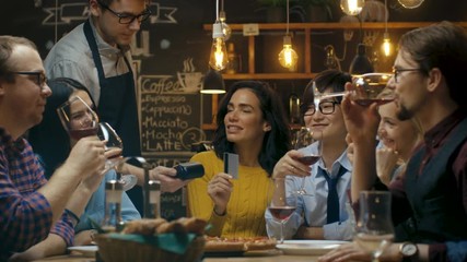 In the Bar Waiter Holds Card Machine and Beautiful Woman Pays for Her Order with Contactless Credit Card. She's Surrounded by Friends They all Have Great Time. 