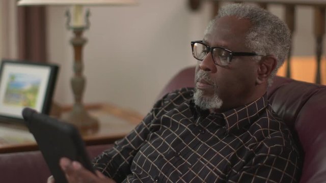 A happy elderly African American grandpa smiling and waving, chatting on a tablet computer. Authentic family feel. Social distancing. Slow motion (48fps) Prores file.