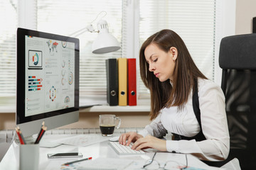 Beautiful smiling brown-hair business woman in suit and glasses sitting at the desk with cup of coffee and mobile phone, working at modern computer with documents in light office, hands on keyboard