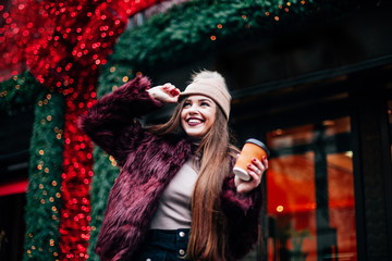 The concept of street fashion. portrait of Young girl dressed in a fashionable outfit. Posing against the window of the boutique girl smiling and drinking coffee