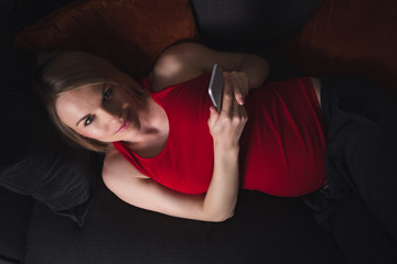 A happy pregnant woman in a red T-shirt and gray pants lies at home on the couch in the evening and holds a mobile phone. A woman is shopping by phone. Online shopping