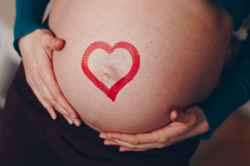 young and beautiful mom to be posing at home with her big baby belly bump. concept of love for unborn child. red heart drawn across the pregnant belly.