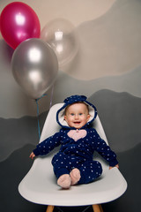Fototapeta na wymiar Funny smiling baby in blue clothes sitting on a white chair with balloons against a background of a gray wall indoor. Baby's first birthday.