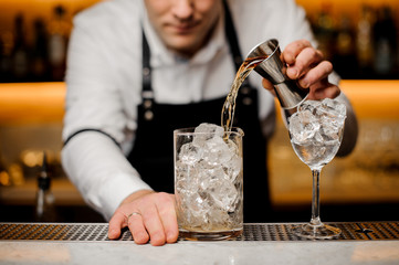 Bartender dressed in a white shirt pouring alcoholic drink into a glass with ice cubes